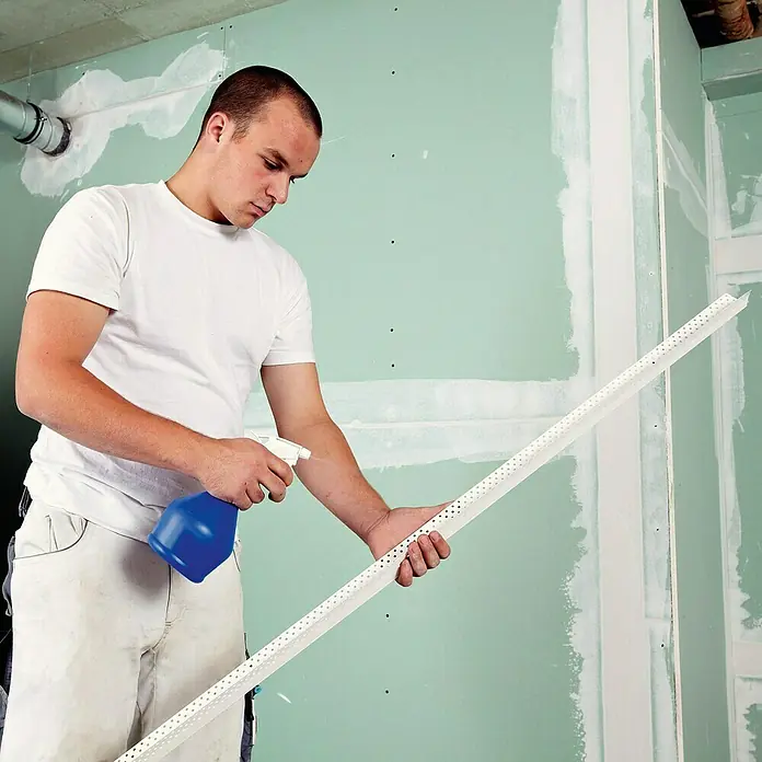 Rigips Kantenschutz für Trockenbau Aqua Bead - 250 cm, Kunststoff - Ein Handwerker befeuchtet eine Putzkante mit einer blauen Sprühflasche vor dem Anbringen an einer grünen Gipskartonwand.
