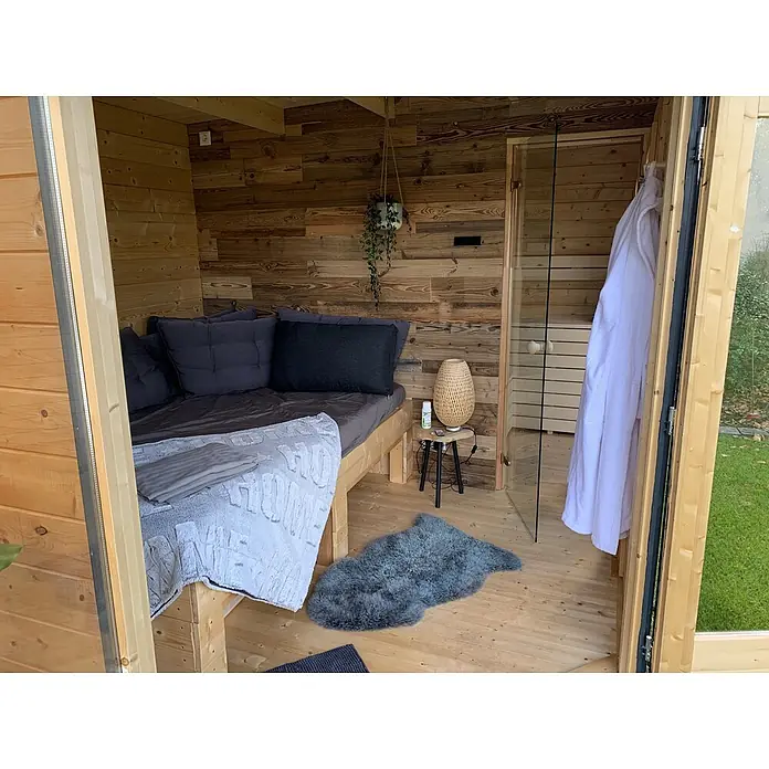 Fjordholz Gartenhaus Birger 44 - Außenmaß inkl. Dachüberstand (B x T): 448 x 298 cm, Holz, ohne Imprägnierung - Gemütliches Holzzimmer mit Bett, dunkelgrauer Bettwäsche, geflochtener Lampe und Sauna durch Glastür sichtbar.