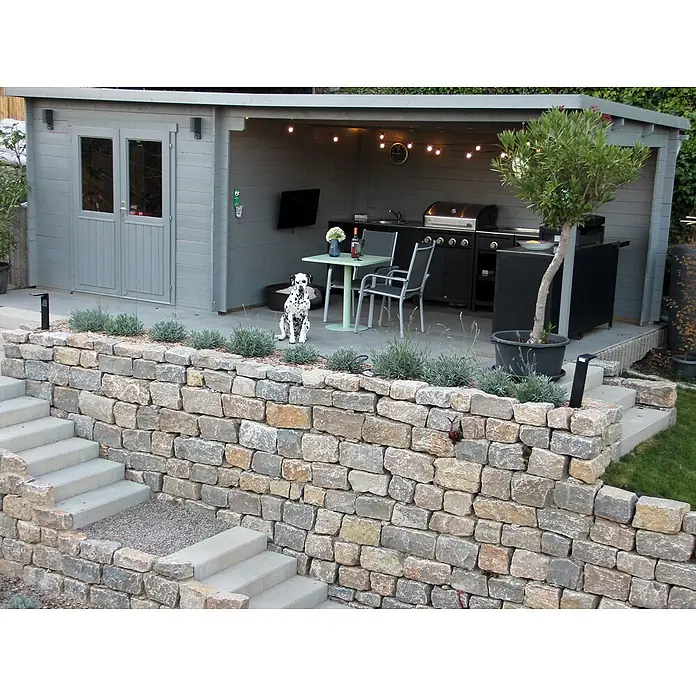 Fjordholz Gartenhaus Gent 28 B - Außenmaß inkl. Dachüberstand (B x T): 691 x 298 cm, Holz, Farbbehandlung Lichtgrau - Ein einladender Outdoor-Unterhaltungsbereich mit grauem Gartenhaus, offener Bar/Küche, Dalmatiner und Steinmauer mit Treppe.