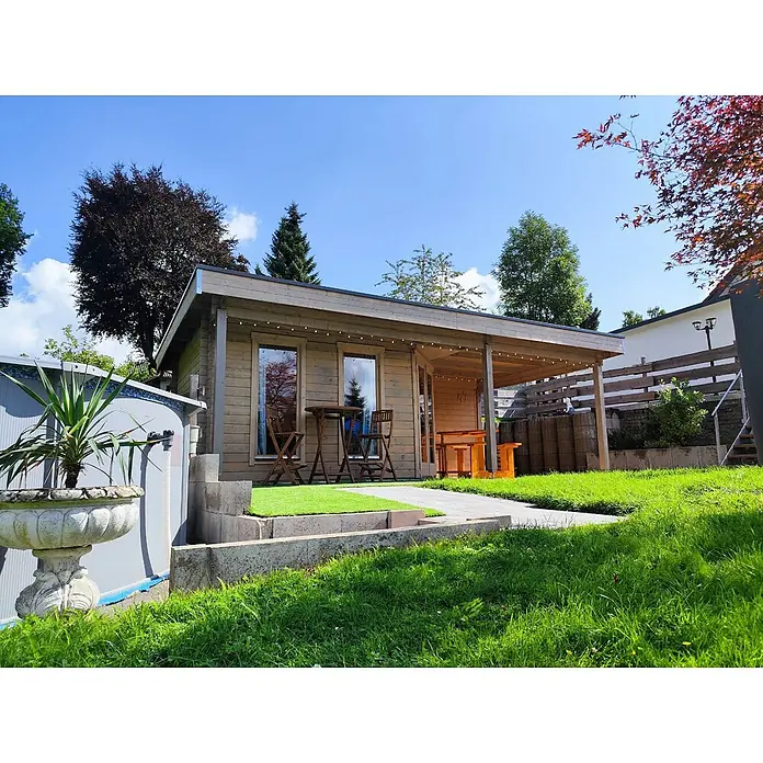 Fjordholz Gartenhaus Nora 44 I Modern - Außenmaß inkl. Dachüberstand (B x T): 725 x 320 cm, Holz, ohne Imprägnierung - Ein helles Holzgartenhaus mit überdachter Terrasse, Fenstern und Sitzgelegenheiten im sonnigen grünen Garten.