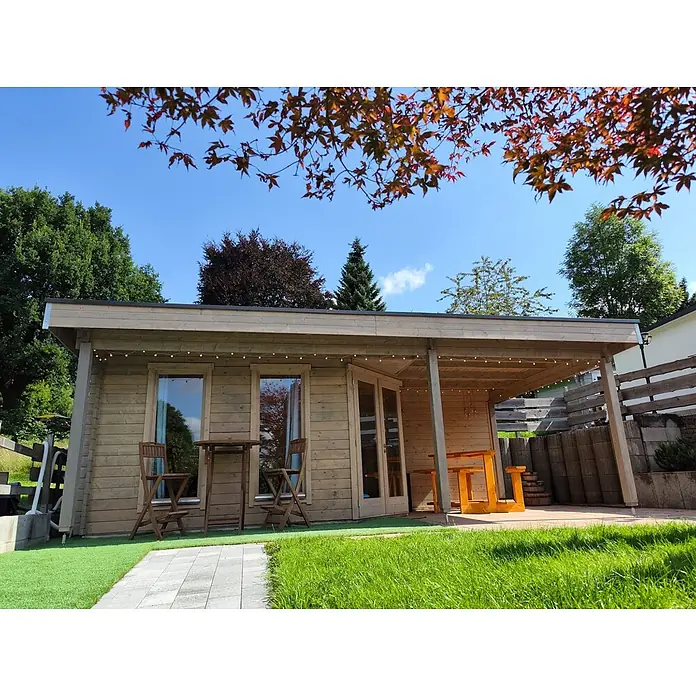 Fjordholz Gartenhaus Nora 44 I Modern - Außenmaß inkl. Dachüberstand (B x T): 725 x 320 cm, Holz, ohne Imprägnierung - Helles Holzgartenhaus mit überdachter Terrasse, ausgestattet mit Gartenmöbeln und Lichterkette, in einem sonnigen Garten.