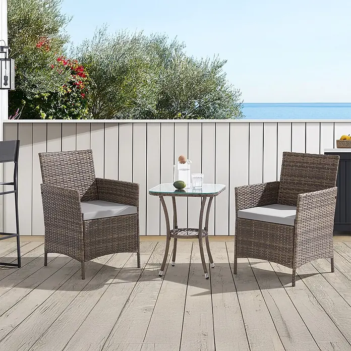 SVITA Loungemöbel-Set - 3 -tlg., Polyrattan, Braun - 3-teiliges braunes Rattan-Terrassen-Set mit grauen Kissen und Glastisch auf einer Holzterrasse mit Meerblick.