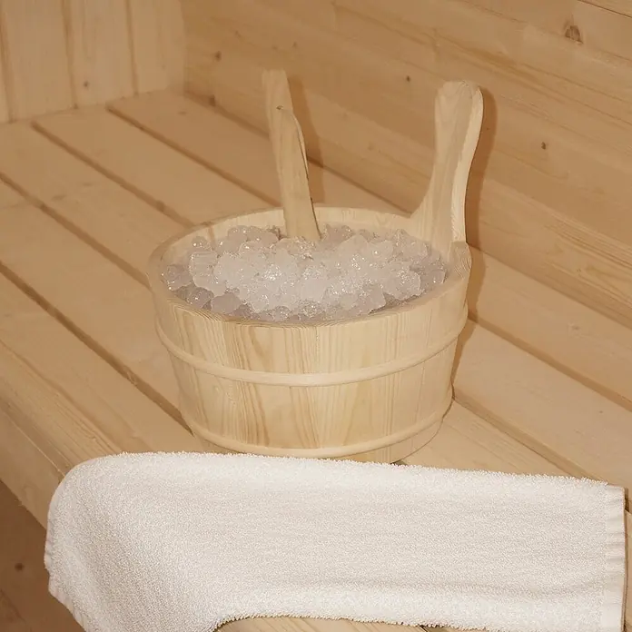 Home Deluxe Fasssauna CUBE L - Elektroofen, Holz - Holzsaunaeimer gefüllt mit Eis und zwei Schaufeln, dazu ein weißes Handtuch auf einer Holzbank.