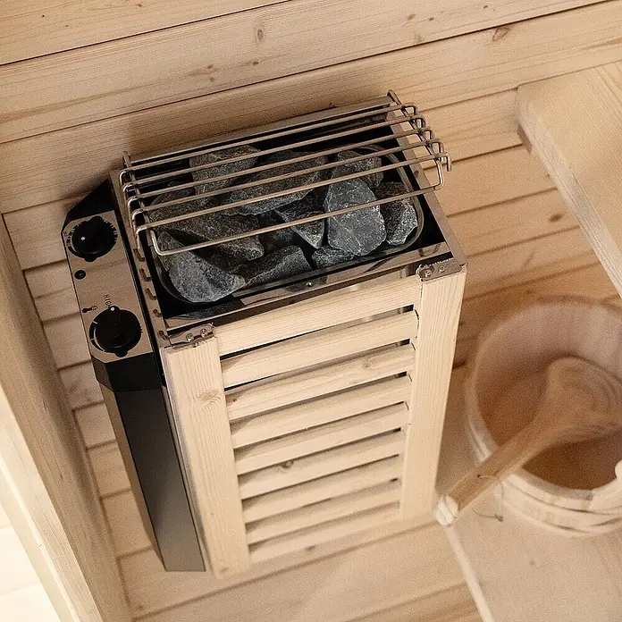 Home Deluxe Fasssauna CUBE S - Elektroofen, Holz - Elektrischer Saunaofen mit grauen Steinen, Edelstahlplatte, schwarzen Bedienelementen und Holzschutzgitter in einer Sauna.