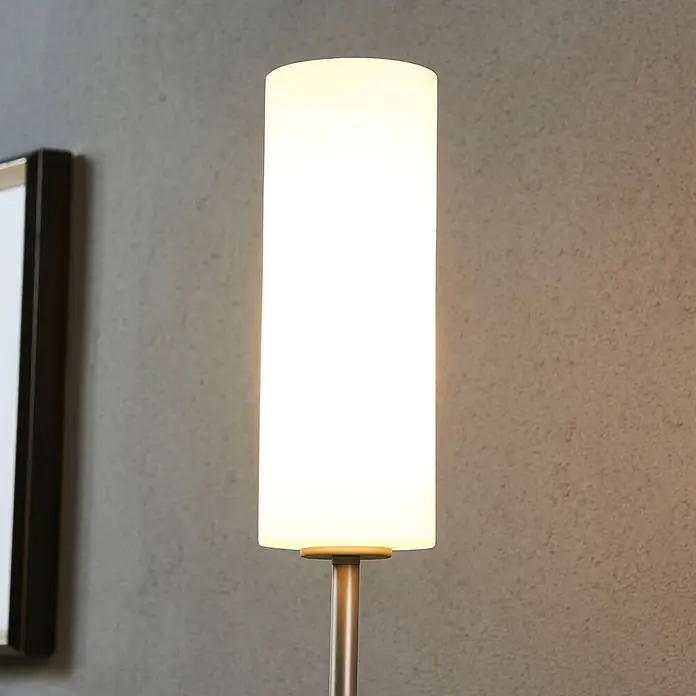 Lindby Stehleuchte Vinsta - alu / grau / zink, weiß / opal, E27 - Leuchtende weiße Zylinderlampe mit Metallsockel vor einer strukturierten Wand.
