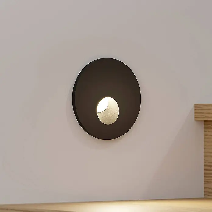Arcchio Wandleuchte Vexi - L x B x H: 7,5 x 7,5 x 4,5 cm, schwarz - Eine dunkle runde Wandleuchte wirft ein warmes halbkreisförmiges Licht nach unten, montiert an einer hellen Wand über einer Holzoberfläche.