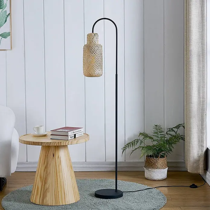 Lindby Stehleuchte Venora - schwarz, Holz hell, E27 - Stehlampe aus schwarzem Metall mit natürlichem Rattan-Schirm, beleuchtet einen gemütlichen Wohnbereich neben einem Holztisch.