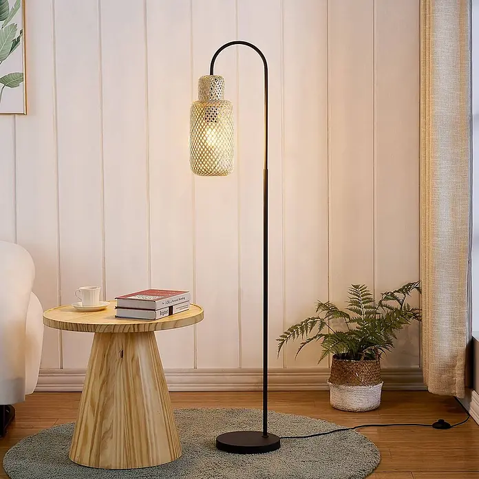 Lindby Stehleuchte Venora - schwarz, Holz hell, E27 - Eine Stehlampe mit schwarzem gebogenem Gestell und gewebtem Naturfaserschirm, die eine gemütliche Wohnzimmerecke beleuchtet.