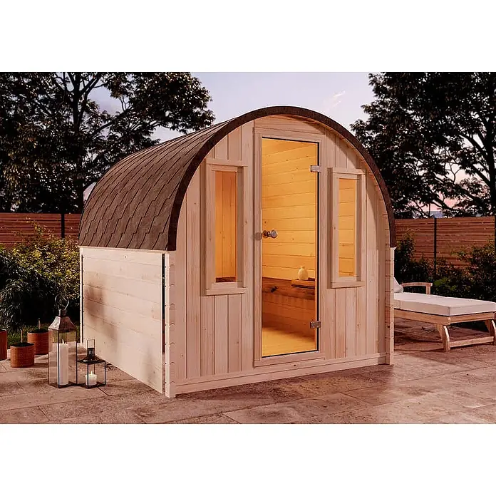 FinnTherm Fasssauna ScandiPod Comfort - Holz, L x B x H: 226 x 21 x 210 cm - Helle Holzfasssauna mit dunklem Schindeldach und Glastür auf einer Terrasse in der Dämmerung. Innenraum beleuchtet, bereit zur Entspannung.