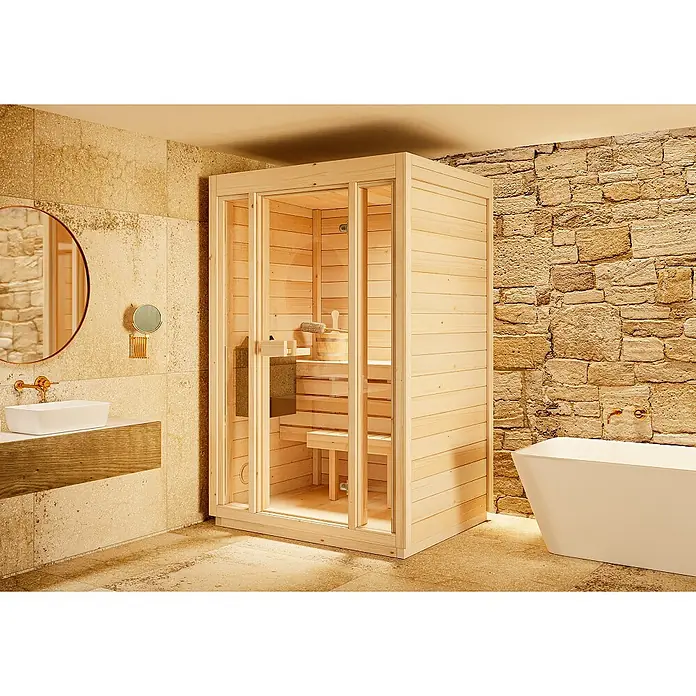 FinnTherm Massivholzsauna Kuuma S - Elektroofen - Helle Holzsaunakabine mit Glastür, in einem modernen Badezimmer mit Steinwänden, Waschbecken und Badewanne.