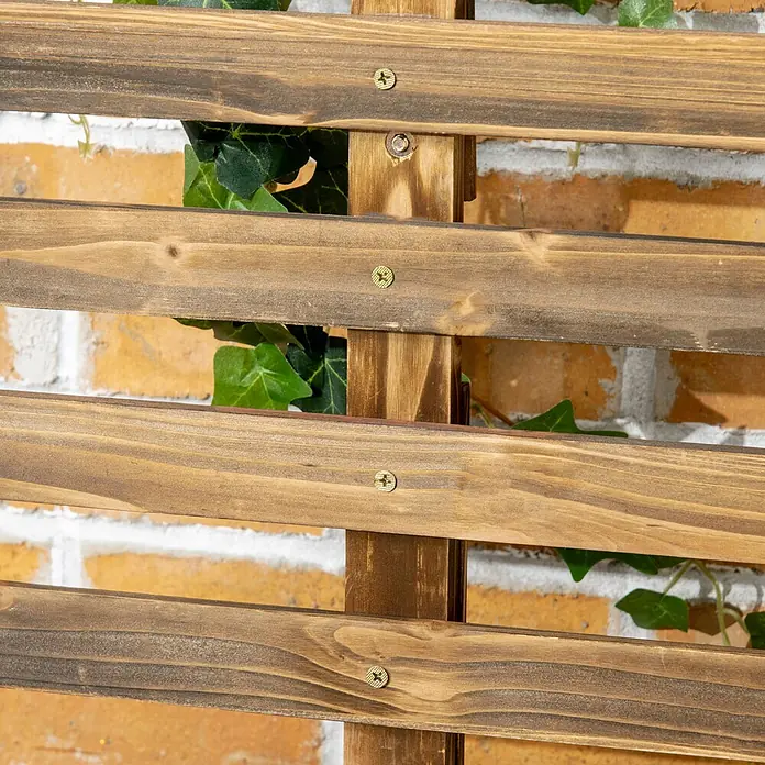 Furnicato Hochbeet - 65 x 31 x 147 cm, Holz, Verkohlt - Braunes Holzspalier mit waagerechten Latten und Messingschrauben, mit grünem Efeu vor einer Ziegelwand.