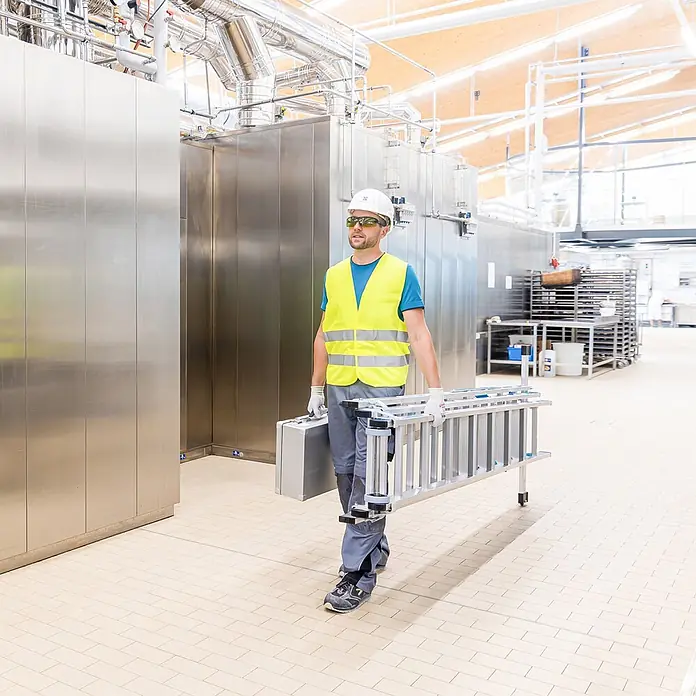 Zarges Mehrzweckleiter Mehrzweckleiter Skymaster Plus X 3x8 Stufen - Anzahl Tritte: 8 Stk., Arbeitshöhe: 6.550 m, Aluminium - Mann trägt silberne Klappleiter und Werkzeugkasten in Fabrik.