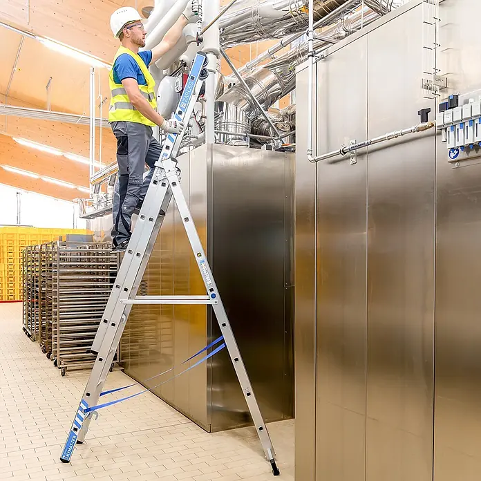 Zarges Mehrzweckleiter Mehrzweckleiter Skymaster Plus X 3x8 Stufen - Anzahl Tritte: 8 Stk., Arbeitshöhe: 6.550 m, Aluminium - Mann auf einer Aluminium-Mehrzweckleiter, der hohe Industriemaschinen erreicht. Die Leiter hat blaue Sicherheitsgurte.