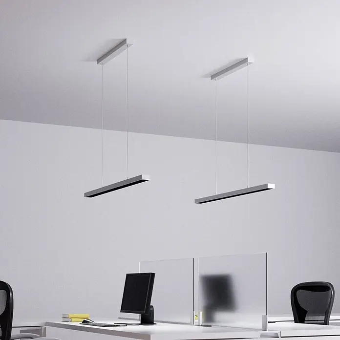Arcchio Pendelleuchte Jolinda - alu / grau / zink, silber (RAL 9006) - Zwei silberne lineare Pendelleuchten beleuchten einen Bürobereich mit Tischen und Monitoren.