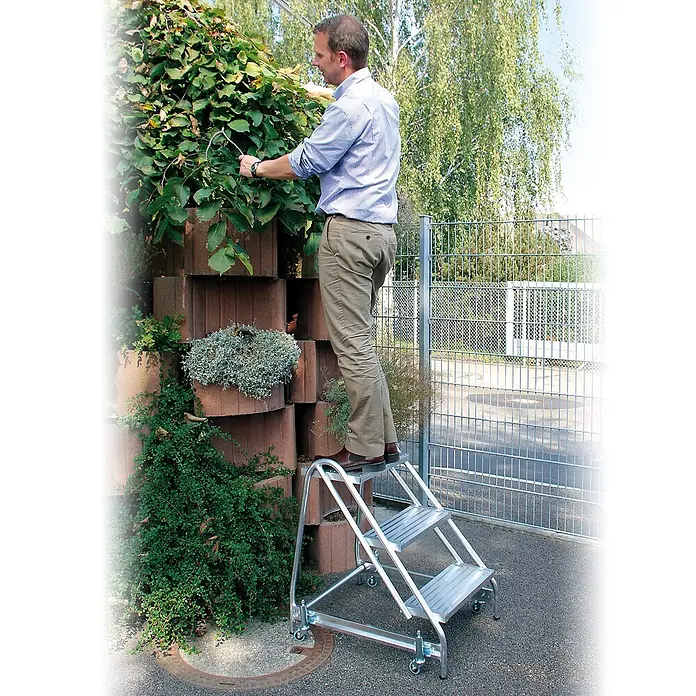 Munk Arbeitsplattform Stahl-Gitterrost Arbeitspodest 2 Stufen - Anzahl Tritte: 2 Stk., Arbeitshöhe: 2.500 m, Aluminium - Mann auf einem silbernen Aluminium-Rollhocker, der eine Hecke schneidet. Der tragbare Hocker hilft ihm, hohe Äste sicher zu erreichen.