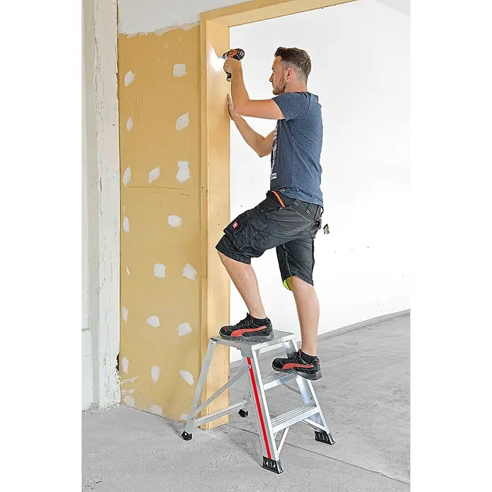 Hymer Arbeitsplattform Klapptritt einseitig begehbar 4 Stufen - Anzahl Tritte: 4 Stk., Arbeitshöhe: 2.840 m, Aluminium - Silberne zweistufige Klappleiter mit roten Akzenten, von einem Mann zum Bohren an einem Türrahmen verwendet.