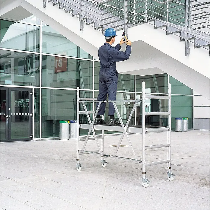 Iller Leiter Rollgerüst Professional Klappgerüst Grundeinheit 0,75x1,80 m | 2,95m AH - Arbeitshöhe: 2.950 m - Arbeiter nutzt ein fahrbares Aluminium-Rollgerüst, um Aufgaben an einer Gebäudefassade auszuführen.