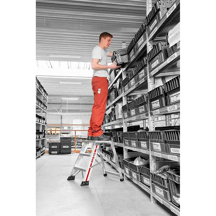 Hymer Arbeitsplattform Klapptritt einseitig begehbar 4 Stufen - Anzahl Tritte: 4 Stk., Arbeitshöhe: 2.840 m, Aluminium - Mann nutzt eine silberne Klapptrittleiter mit roten Akzenten, um Artikel im Lager zu erreichen.