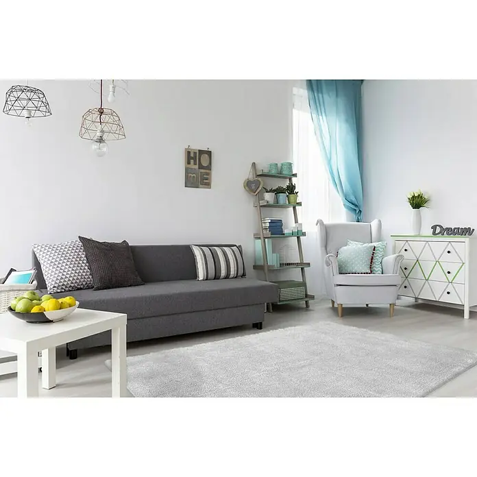 Kayoom Hochflorteppich Bali - Silbergrau, 150 x 80 cm, 100 % Polyester - Wohnzimmer mit grauer Couch, weißem Tisch und Teppich, hellblauem Vorhang und weißen Möbeln. Dekorative Elemente und Lampen runden das Bild ab.