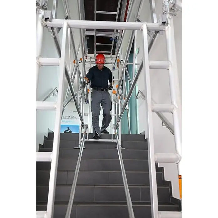 Layher Rollgerüst Treppen-Kit für Fahrgerüst Uni Standard P2 mit 2 Durchgängen 7,73m AH - Arbeitshöhe: 7.730 m - Arbeiter auf modularem Aluminium-Treppengerüst in einem Gebäude. Die silberne Struktur bietet temporären Zugang über dunkle Treppen.