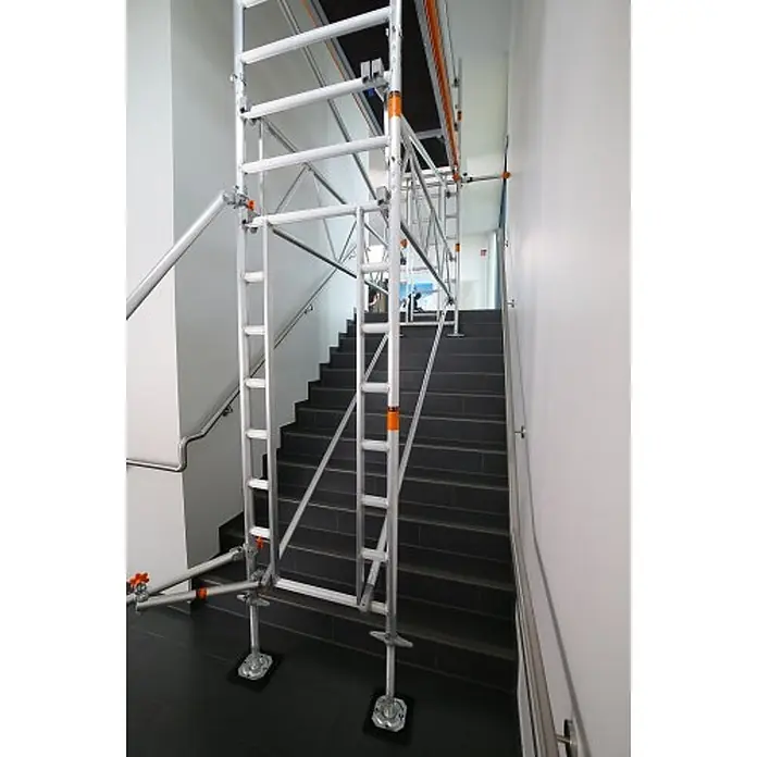 Layher Rollgerüst Treppen-Kit für Fahrgerüst Uni Standard P2 mit 2 Durchgängen 7,73m AH - Arbeitshöhe: 7.730 m - Silbernes Aluminium-Treppenhausgerüst mit orangefarbenen Akzenten, sicher über einer dunklen Treppe aufgebaut.