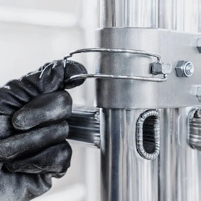 Werner Rollgerüst Tower Klappgerüst 7,00m AH - Arbeitshöhe: 6.900 m - Eine behandschuhte Hand sichert eine Drahtklammer an einem silbernen Metallteil eines modularen Regalsystems.