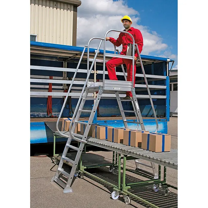 Munk Treppenpodest Überbrückung beidseitig begehbar 7 Stufen -  - Silberne Aluminium-Arbeitsplattform mit Handläufen, von einem Arbeiter zum Erreichen eines Lkw genutzt, neben einem Förderband mit Kartons.