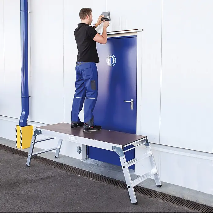 Munk Arbeitsplattform Arbeitsdiele klappbar - Anzahl Tritte: 3 Stk., Arbeitshöhe: 2.690 m, Aluminium - Mann auf einer Arbeitsplattform aus Aluminium mit brauner Oberfläche installiert etwas über einer Tür. Bietet eine stabile erhöhte Arbeitsfläche.