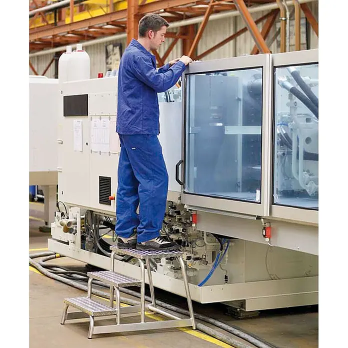 Zarges Arbeitsplattform Aluminium Montagetritt starr AFA P 1 Stufe - Anzahl Tritte: 1 Stk., Arbeitshöhe: 2.200 m, Aluminium - Mann nutzt einen silbernen dreistufigen Metall-Trittschemel mit Riffelblech, um eine Maschine in einer Fabrik zu erreichen.