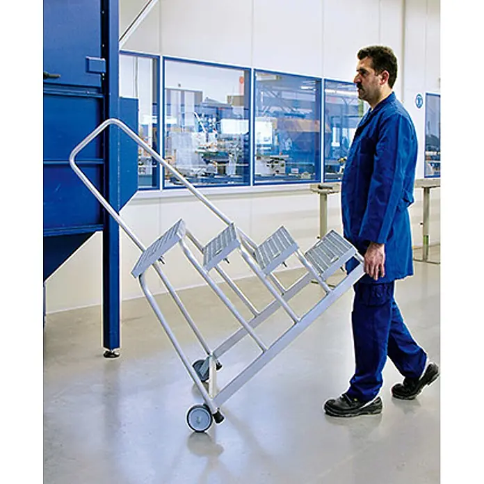 Munk Arbeitsplattform Arbeitspodest fahrbar mit Stahl-Gitterrost mit Handlauf 5 Stufen - Anzahl Tritte: 5 Stk., Arbeitshöhe: 3.000 m, Aluminium - Graue mobile Stehleiter mit drei geriffelten Stufen und Rollen, von einem Arbeiter gezogen.