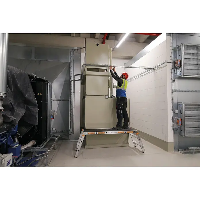 Layher Arbeitsplattform Arbeitsplattform TOPIC 1065 klappbar - Anzahl Tritte: 3 Stk., Arbeitshöhe: 2.650 m, Aluminium - Arbeiter auf einer silber-orangen Layher Arbeitsplattform, der Industrieanlagen in einer Halle justiert.