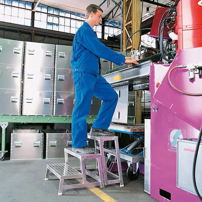 Zarges Arbeitsplattform Aluminium-Arbeitspodest AMA P Modul A - Arbeitshöhe: 2.200 m, Aluminium - Mann nutzt eine mehrstufige Metallplattform mit Riffelblech, um eine Maschine in einer Industrieumgebung zu bedienen.