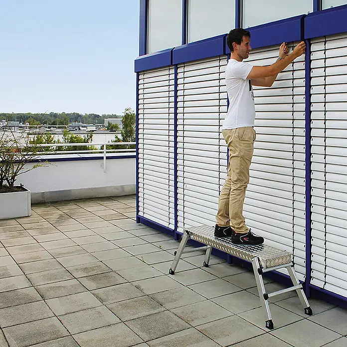 Munk Arbeitsplattform Aluminium Arbeitsplattform mit Klappfüßen - Anzahl Tritte: 2 Stk., Arbeitshöhe: 2.500 m, Aluminium - Aluminium-Arbeitsplattform mit Riffelblech-Oberfläche, von einem Mann genutzt, um Jalousien an einer Fassade zu erreichen.