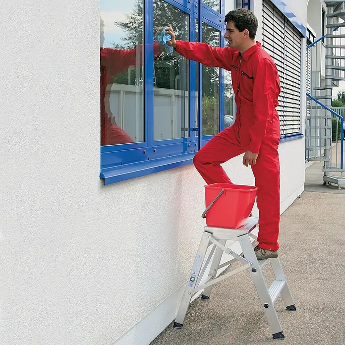 Munk Arbeitsplattform Aluminium Arbeitspodest klappbar 4 Stufen - Anzahl Tritte: 4 Stk., Arbeitshöhe: 3.000 m, Aluminium - Silberner Tritthocker mit rotem Eimer, auf dem ein Mann Fenster putzt.