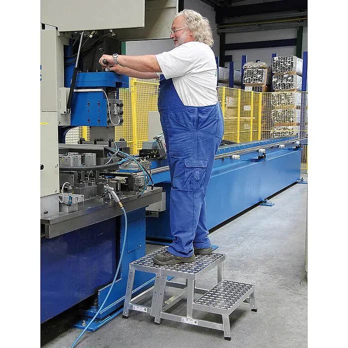 Munk Arbeitsplattform modulares Arbeitspodest Erweiterungsmodul 1 - Arbeitshöhe: 2.400 m, Aluminium - Silberner zweistufiger Industrietritt mit Riffelblech-Trittflächen, von einem Mann in einer Fabrik genutzt.