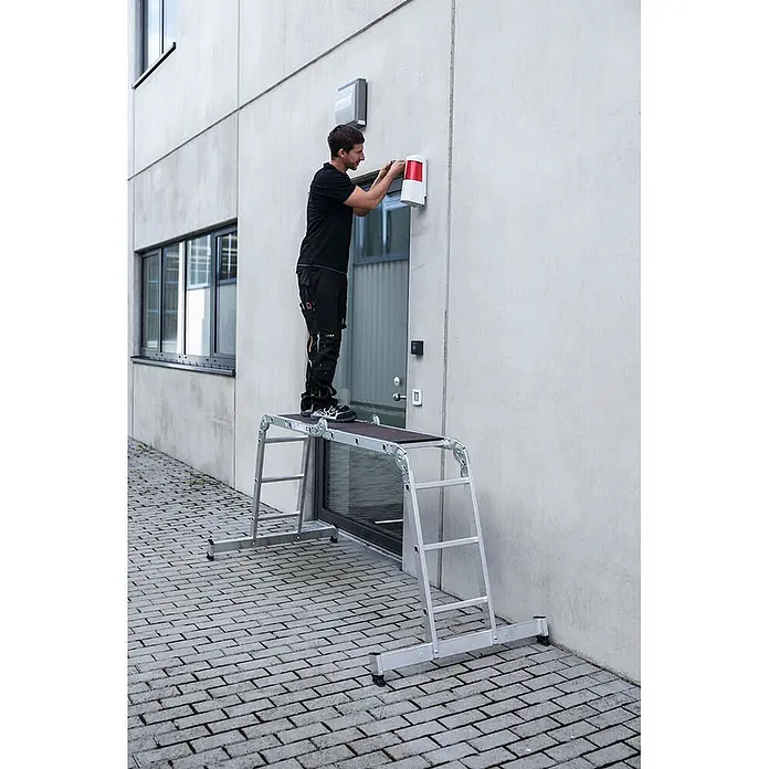 Munk Mehrzweckleiter Vielzweckleiter mit Nivello® Traverse und Holzbelag 2x2 + 2x3 Sprossen - Arbeitshöhe: 4.000 m, Aluminium - Mann steht auf einer silbernen Aluminium-Arbeitsplattform mit schwarzer Oberfläche und arbeitet an einer Wand.