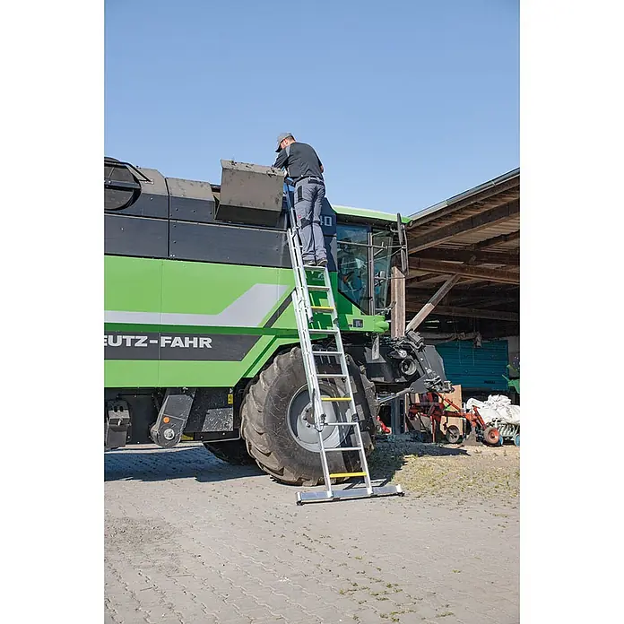 Munk Mehrzweckleiter Stufen- Mehrzweckleiter mit Wandlaufrollen, clipstep R13 und Nivello® Trav - Anzahl Tritte: 8 Stk., Arbeitshöhe: 6.900 m, Aluminium - Eine Aluminium-Sicherheitszugangsleiter mit gelben Stufen lehnt an einer großen grünen Landmaschine, ein Mann steht darauf.