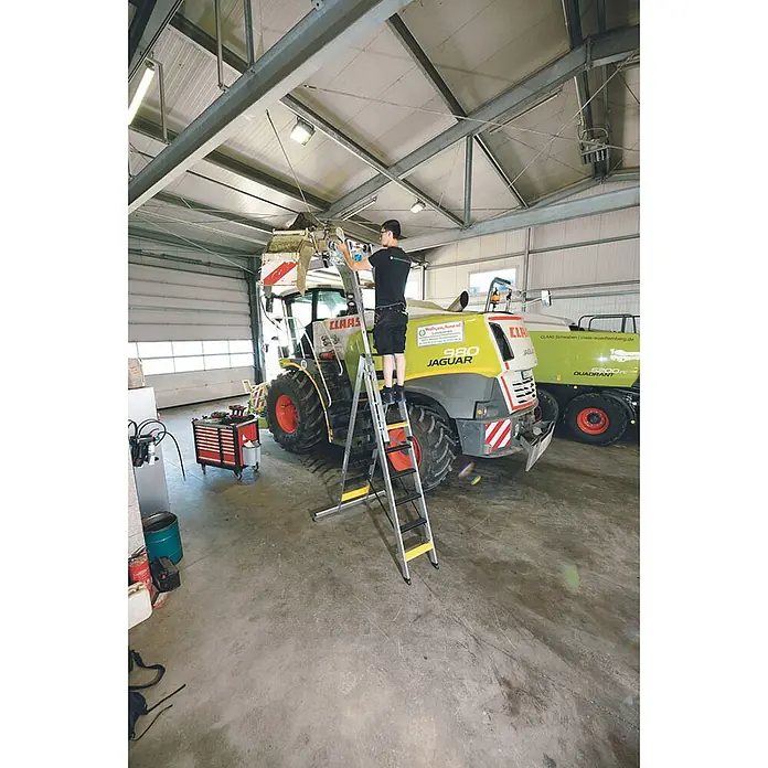 Munk Mehrzweckleiter Stufen- Mehrzweckleiter mit Wandlaufrollen, clipstep R13 und Nivello® Trav - Anzahl Tritte: 8 Stk., Arbeitshöhe: 6.900 m, Aluminium - Aluminium-Klappleiter mit breiten Stufen, wird von einem Mann zur Arbeit an einer großen CLAAS-Landmaschine in einer Werkstatt verwendet.