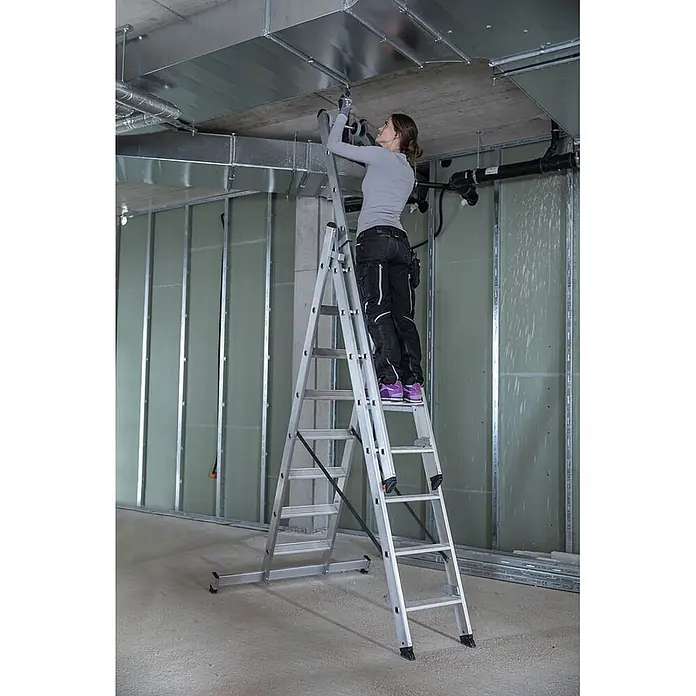 Munk Mehrzweckleiter Stufen- Mehrzweckleiter mit Wandlaufrollen, clipstep R13 und Nivello® Trav - Anzahl Tritte: 8 Stk., Arbeitshöhe: 6.900 m, Aluminium - Silberne Aluminium-Mehrzweckleiter, die von einer Frau für Überkopfarbeiten an Lüftungsrohren in einer Bauumgebung verwendet wird.