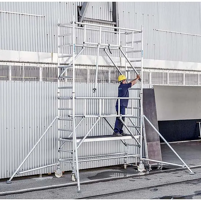 Zarges Rollgerüst Fahrgerüst MultiTower S-PLUS 2T mit Auslegern, doppelte Plattformbreite 2, - Arbeitshöhe: 12.350 m - Silbernes Aluminium-Rollgerüst wird von einem Arbeiter neben einem Gebäude montiert.
