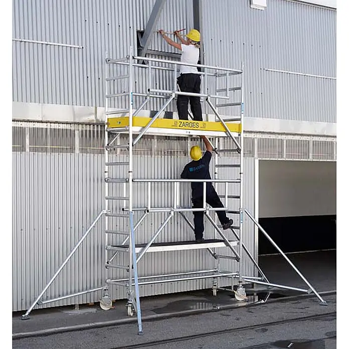 Zarges Rollgerüst Fahrgerüst MultiTower S-PLUS 2T mit Auslegern, doppelte Plattformbreite 2, - Arbeitshöhe: 12.350 m - ZARGES Aluminium-Rollgerüst mit zwei Arbeitern auf Plattformen neben einem Gebäude. Gelbes Sicherheitsbranding.