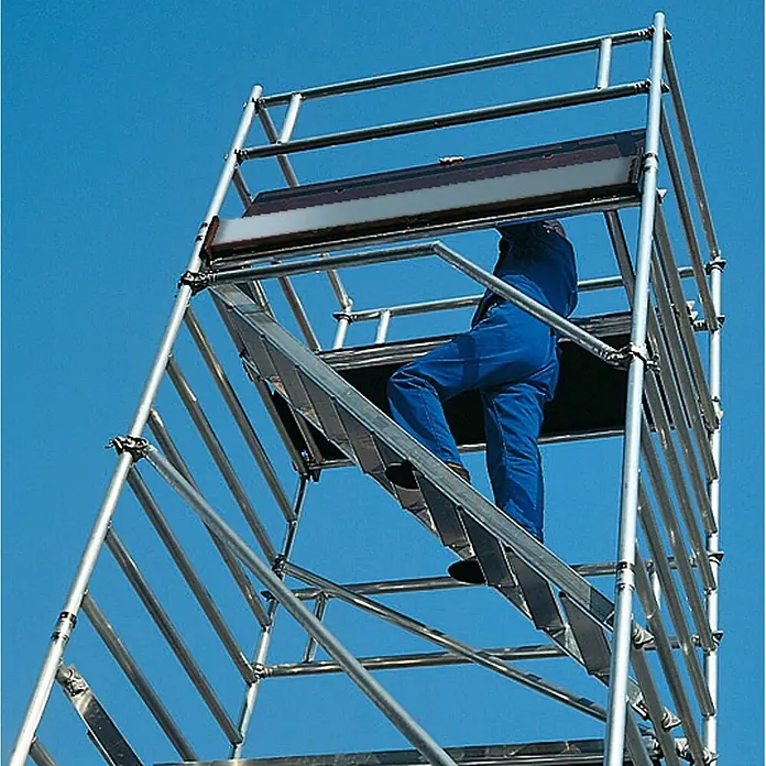 Munk Rollgerüst Rollgerüst mit Schrägaufstiegen 1,35x2,45m Plattform, 10,35m AH - Arbeitshöhe: 10.350 m - Person in blauem Overall erklimmt die Innentreppe eines mobilen Gerüstturms aus silbernem Aluminium vor blauem Himmel.