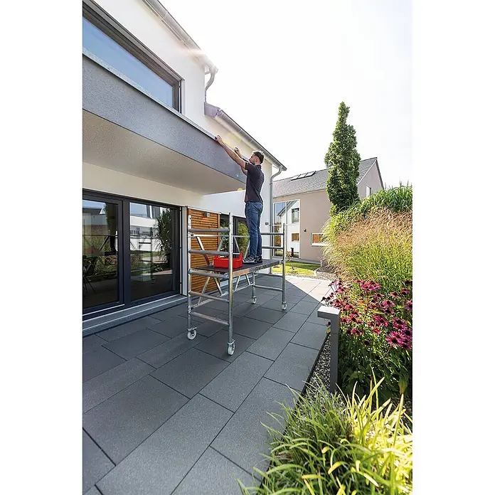 Munk Rollgerüst Klappgerüst, türengängig 75cm Breite 3,00m AH - Arbeitshöhe: 3.000 m - Silbernes Aluminium-Rollgerüst mit Rollen, von einem Mann für Außenarbeiten am Haus genutzt.