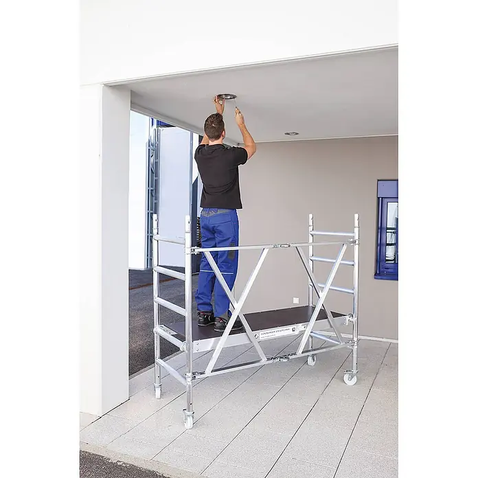 Munk Rollgerüst Klappgerüst, türengängig 75cm Breite 3,00m AH - Arbeitshöhe: 3.000 m - Ein Mann nutzt ein silbernes Aluminium-Rollgerüst mit dunkler Plattform zur Deckenleuchteninstallation.