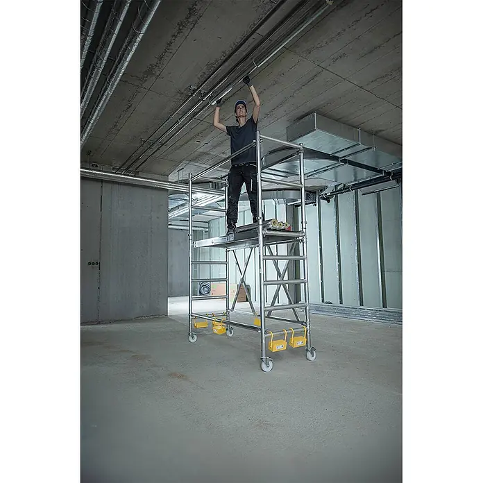 Munk Rollgerüst Klappgerüst mit Ausbaueinheit 75cm Breite 3,60m AH - Arbeitshöhe: 3.600 m - Mobiles Aluminium-Rollgerüst mit gelben Stabilisatoren, von einem Arbeiter für Deckeninstallationen in einem Gebäude verwendet.