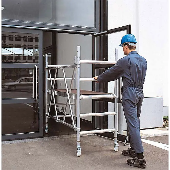 Zarges Rollgerüst Kleingerüst CompactMaster 1T 2,95m AH (DIN EN 1004:2004) - Arbeitshöhe: 2.950 m - Silbernes Aluminium-Rollgerüst auf Rädern wird von einem Arbeiter durch eine Tür geschoben.