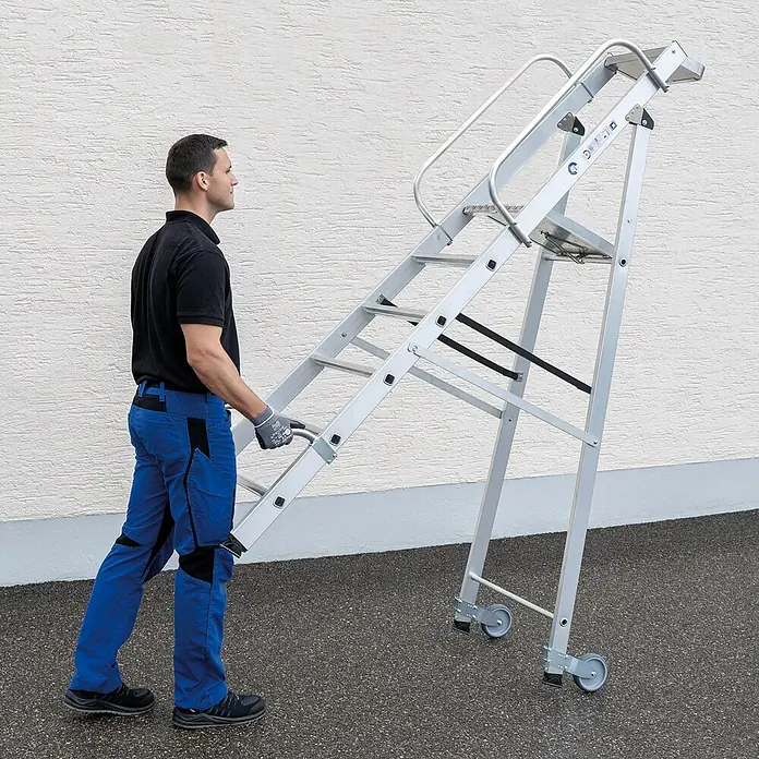Munk Stehleiter Aluminium-Stehleiter mit Rollen und Griff 6 Stufen - Anzahl Tritte: 6 Stk., Arbeitshöhe: 3.400 m, Aluminium - Mann bewegt mobile Arbeitsplattform aus silbernem Aluminium mit Handläufen und Transporträdern.