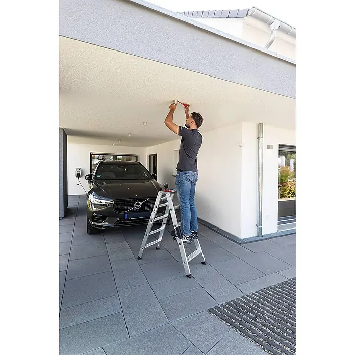 Munk Stehleiter Stehleiter mit ergo-pad beidseitig begehbar 2x5 Stufen - Anzahl Tritte: 5 Stk., Arbeitshöhe: 2.650 m, Aluminium - Mann benutzt eine silberne Aluminium-Klappleiter, um eine Deckenleuchte im Carport zu installieren.