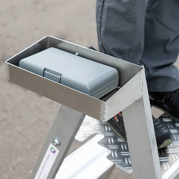 Munk Stehleiter Klapptritt beidseitig begehbar Alu natur 3 Stufen 2,73m AH - Anzahl Tritte: 3 Stk., Arbeitshöhe: 2.730 m, Aluminium - Metall-Werkzeugablage an einer Leiter, hält einen grauen Koffer. Beine einer Person sind auf den Stufen sichtbar.