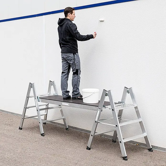 Munk Stehleiter Klapptritt beidseitig begehbar Alu natur 4 Stufen 2,99m AH - Anzahl Tritte: 4 Stk., Arbeitshöhe: 2.990 m, Aluminium - Aluminium-Arbeitsplattform, die einen Mann beim Wände streichen unterstützt, mit einem weißen Eimer auf der Plattform.